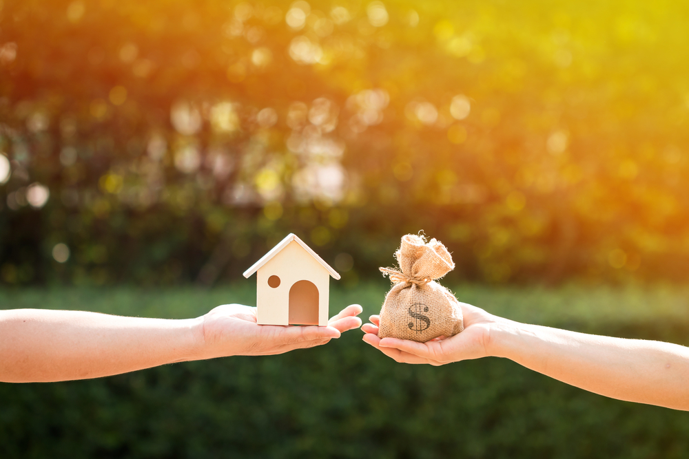 Two people holding a miniature wooden house and a small bag with a dollar sign on it.