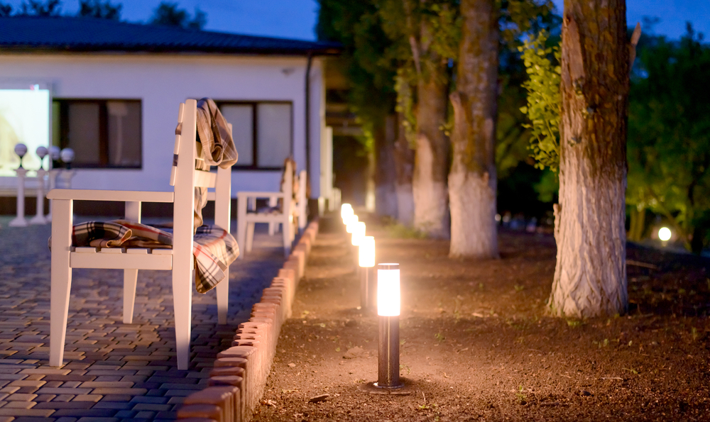 Row of outdoor patio lighting next to a white bench and a row of trees in the evening.