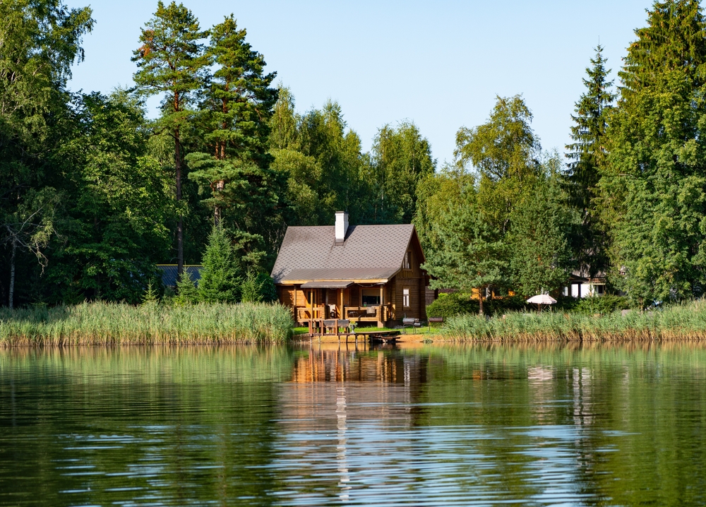 Small, wooden cabin next to a lake surrounded by green trees.