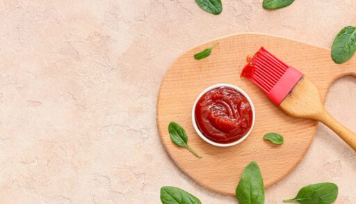 Overhead view of a bowl with barbecue sauce next to a pink silicone brush surrounded by basil leaves.