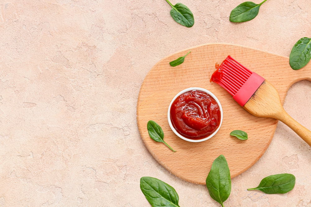 Overhead view of a bowl with barbecue sauce next to a pink silicone brush surrounded by basil leaves.