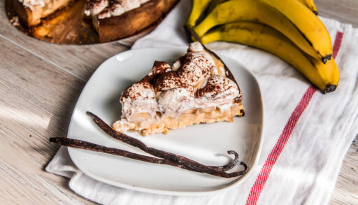 Piece of banofee pie on a white plate with two vanilla beans and a bunch of bananas.