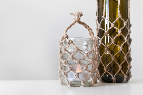 A clear glass jar wrapped in rope with a tea light next to a taller, green bottle.
