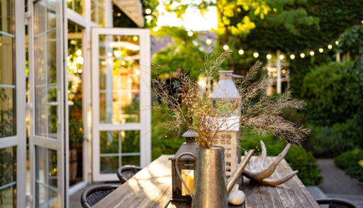 Outdoor dining table with a lantern and metal watering can in the centre and lights hanging overhead.