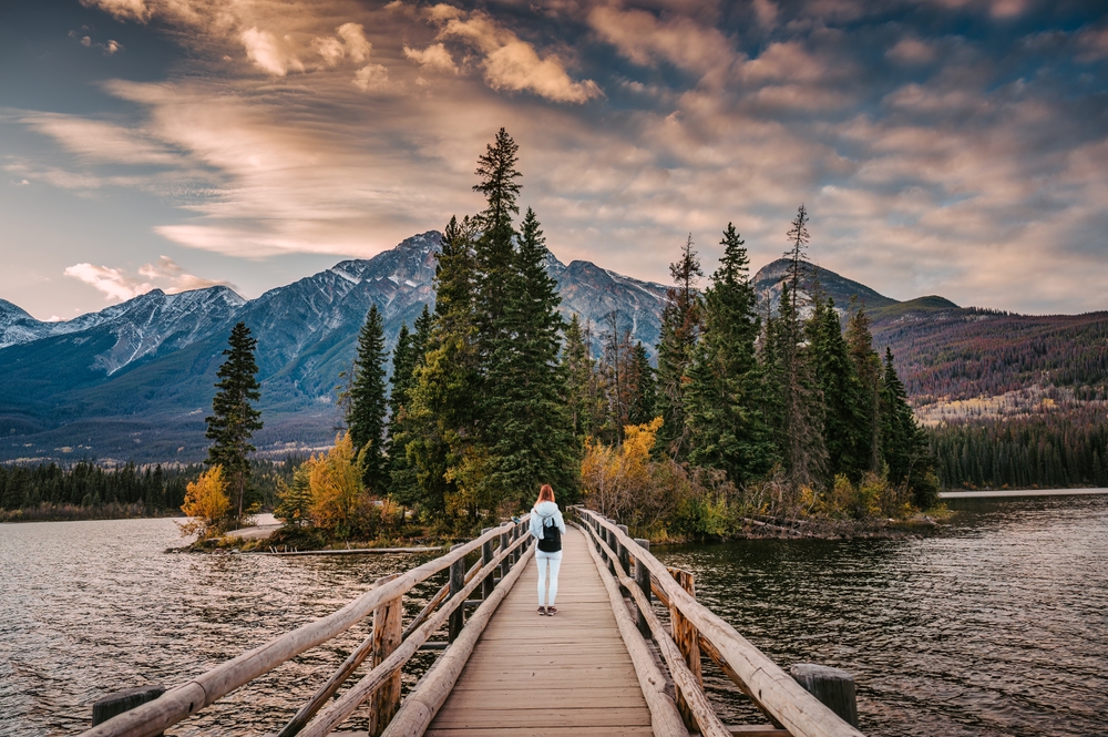 Female tourist standing on a wooden bridge over a lake leading to an island with yellow and pine trees with a mountain in the background.