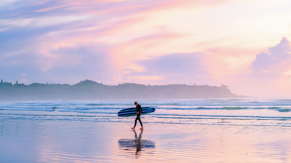 A person walking along Tofino Beach, B.C. with a surfboard at sunset.