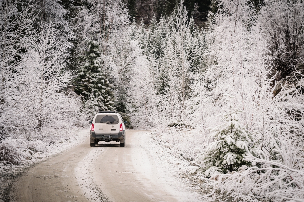White car driving down a snow covered road lined with snowy trees.