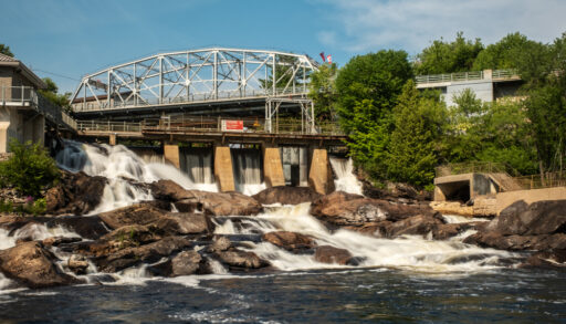Bridge nestled in some trees with water rushing through a dam underneath.