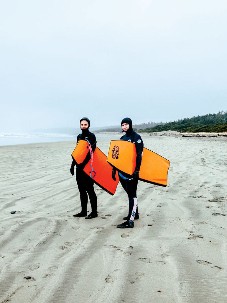 two women wearing full wetsuits carrying surfboards on a beach