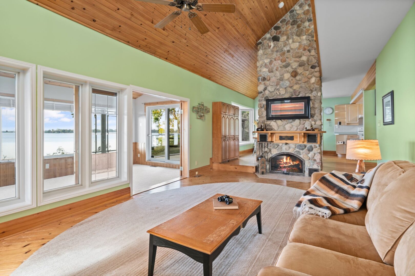 A bright living area with a couch, a wood-panelled ceiling, and a floor-to-ceiling stone fireplace. There is a sunroom just off the living room.