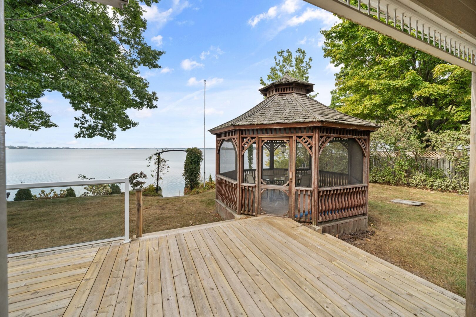 A gazebo sits on the edge of a large back deck of a cottage, with views of the lake.