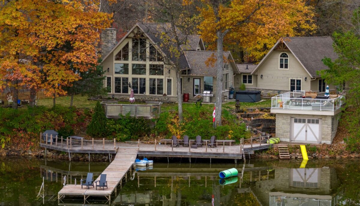 A large cottage with huge windows, a boathouse, and a long dock sits on the shore of a calm lake, surrounded by trees with colourful fall leaves.