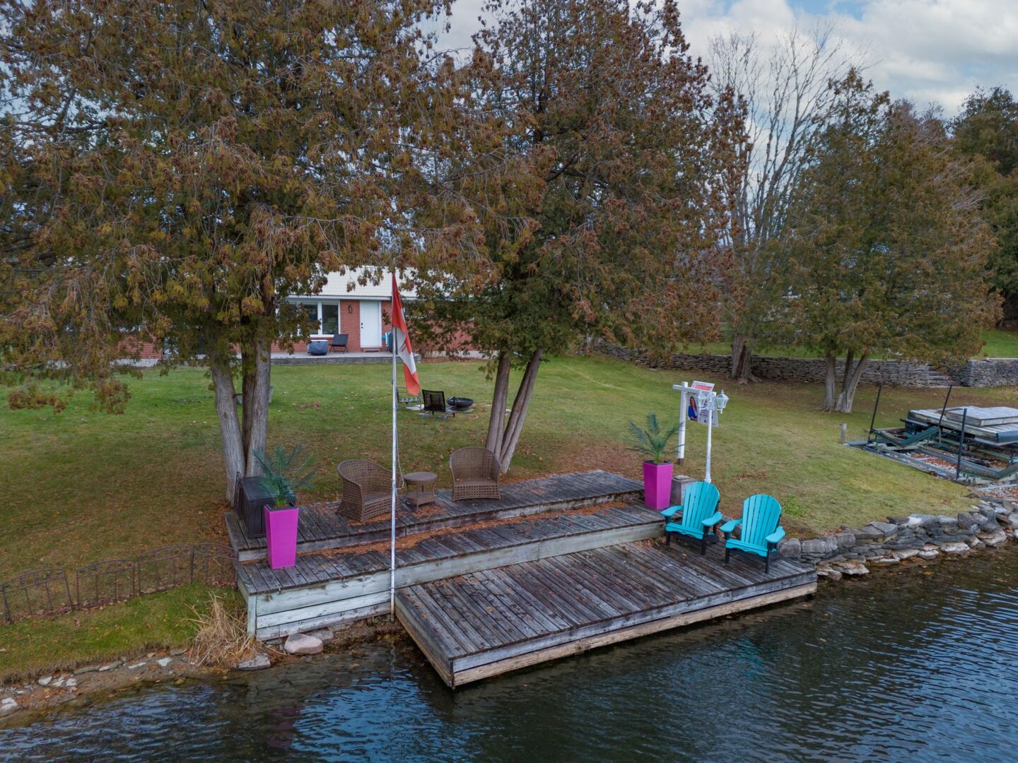 A tiered wooden deck with three levels leads from a grassy yard down to a river. Pink planters and blue Muskoka chairs sit on the deck.