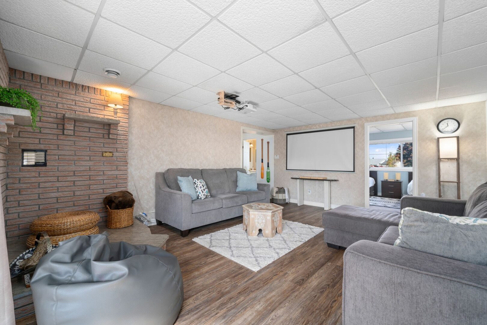A wide living area with grey couches, wood-panelled flooring, and a projector mounted to the ceiling, which is facing a projector screen on one of the walls.