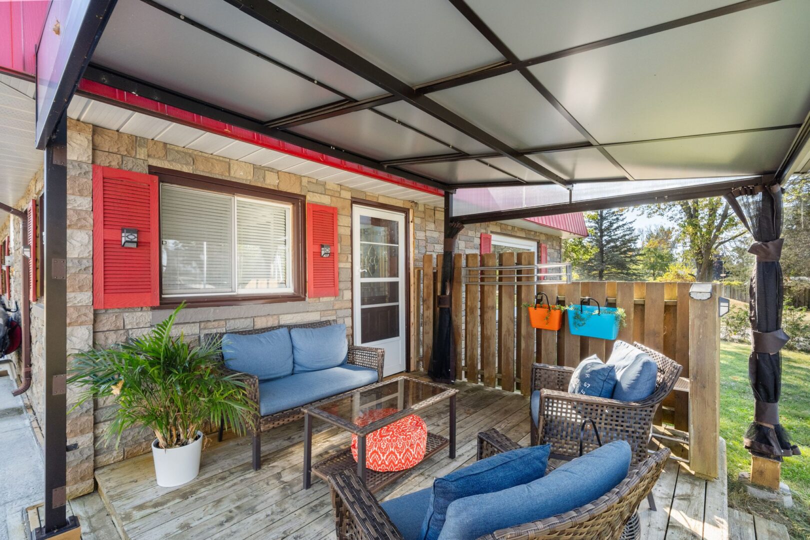 A covered deck with outdoor seating extends off the back of a bungalow with red window trim.