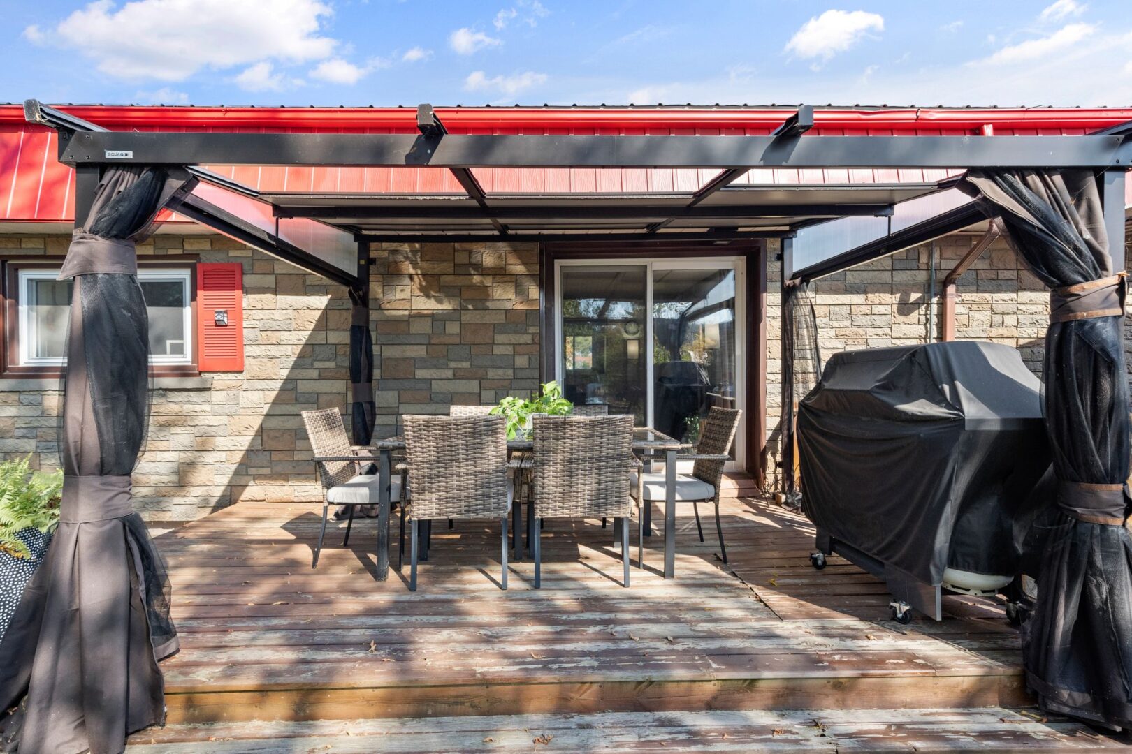 A covered deck extends off the side of a bungalow. There is outdoor dining furniture and a barbecue on the deck.
