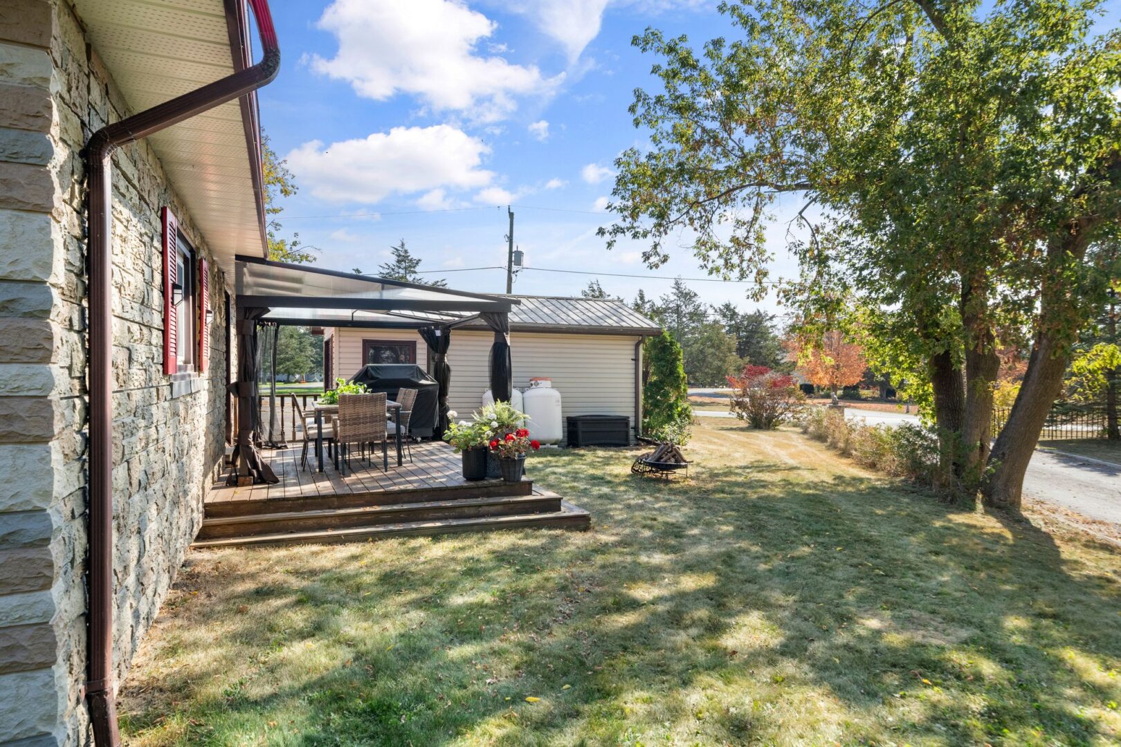 A shady, grassy yard beside a bungalow. A covered deck extends off one side of the home.