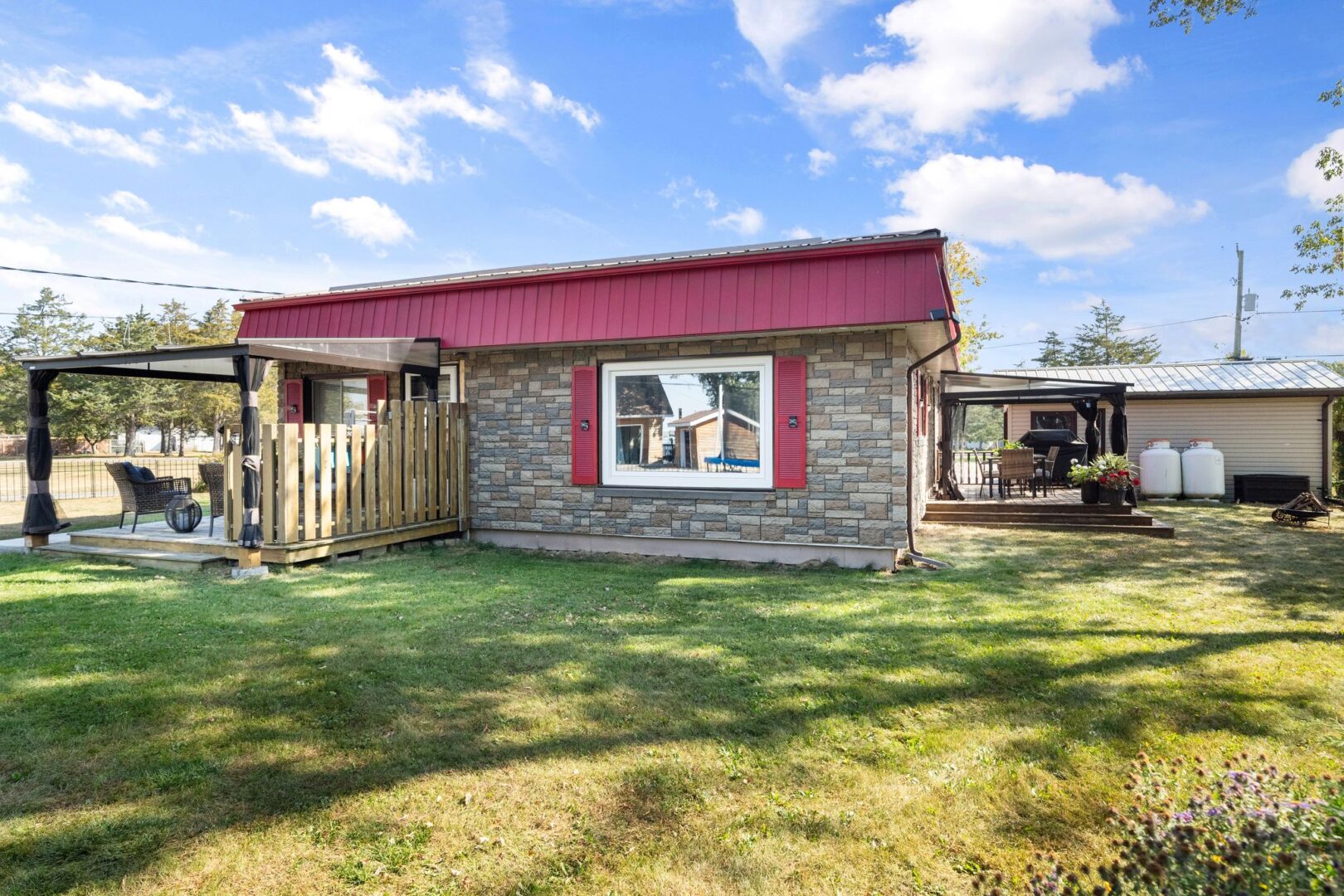 A bungalow with red roof and window trim sits on a wide grassy lot. Two decks extend off adjacent sides of the home.