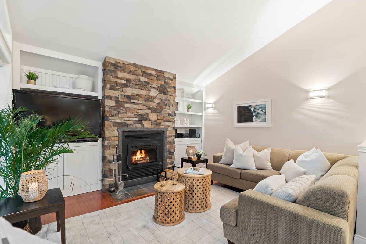A cozy cottage living space with two couches, an area rug, and a wood-burning fireplace framed in floor-to-ceiling stone.