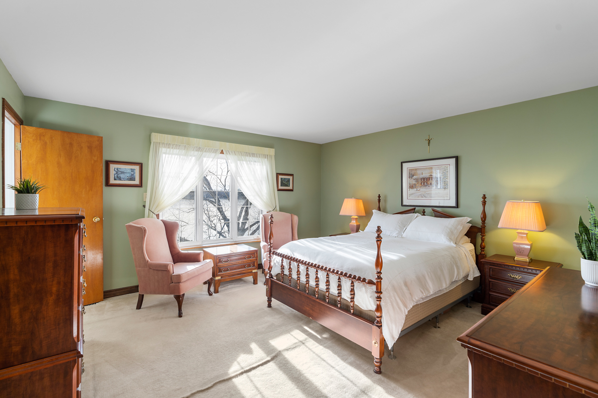 A large, bright bedroom with a big bed, light green walls, carpet flooring, and two light pink armchairs.