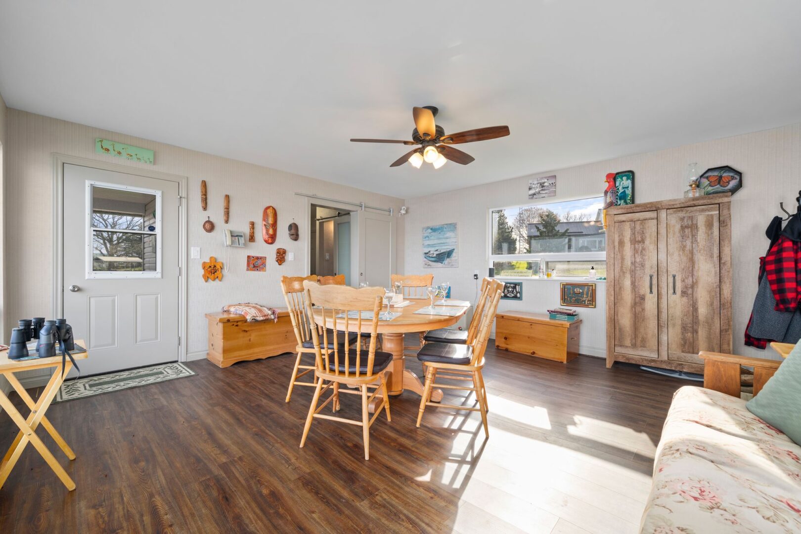 A bright interior space of a waterfront bunkie, with wood flooring, a table and chairs, a couch, and a ceiling fan.