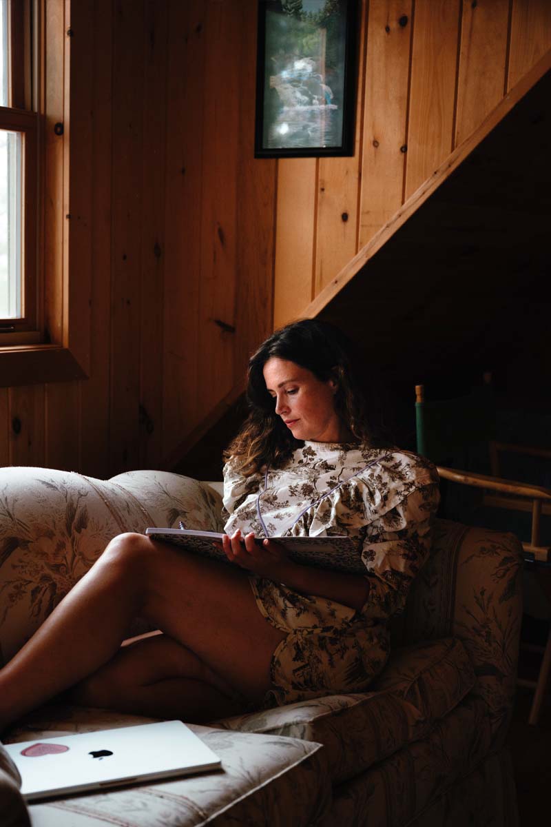 Carley Fortune, pictured, has a book open in her arms while she sits on a couch next to a window at her cottage.