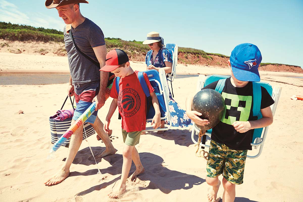 A family carries beach chairs and picnic bags along a beach