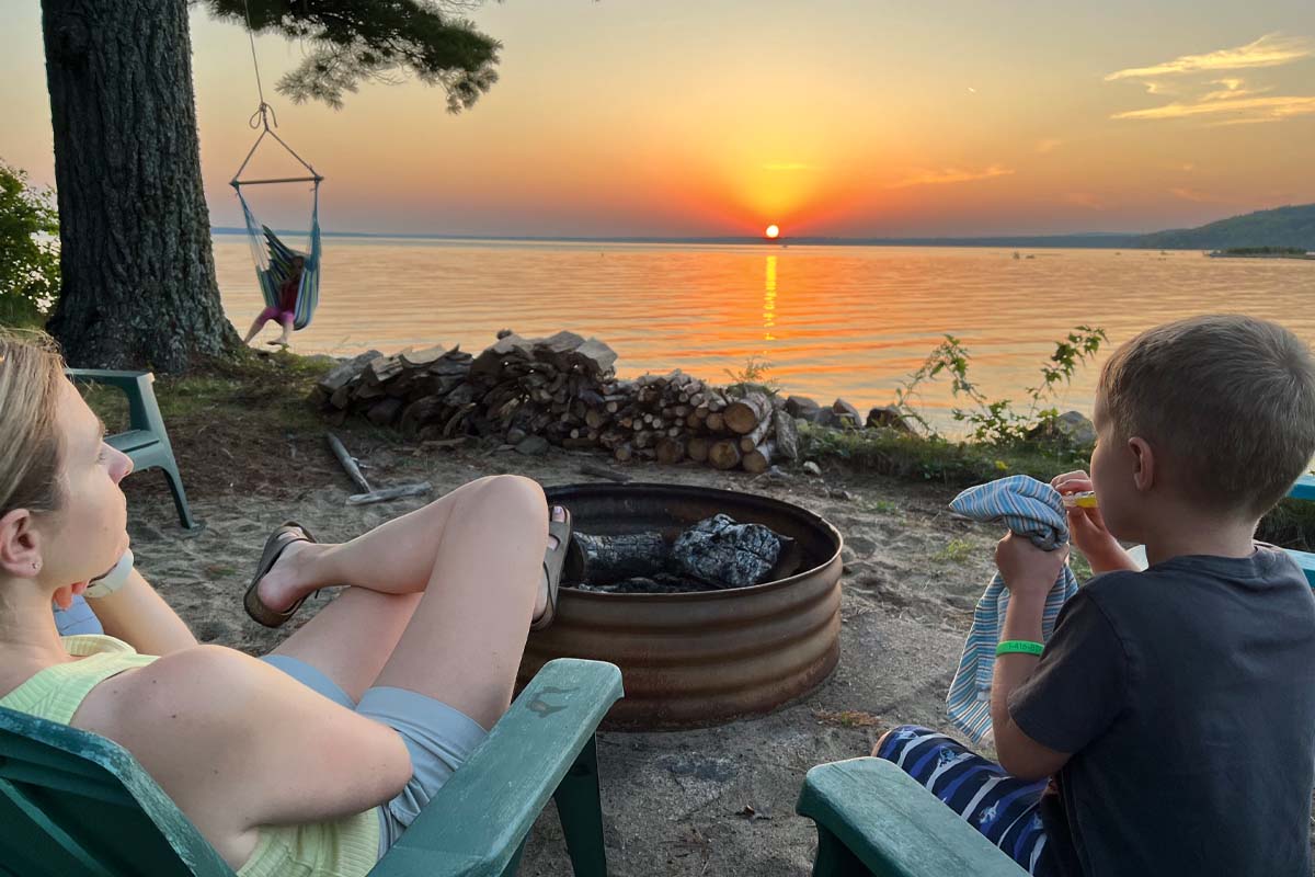 Kids sit around a firepit beside a lake with the sun setting over the horizon.