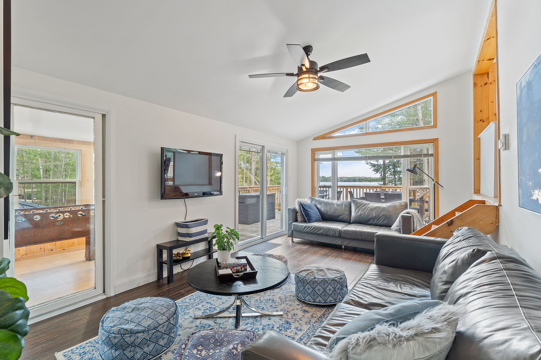 A living area with lots of seating. Big windows look out to a deck and show a sliver of lake beyond. Two sets of sliding glass doors lead out of the room; one into a sunroom, and the other out to a big deck.