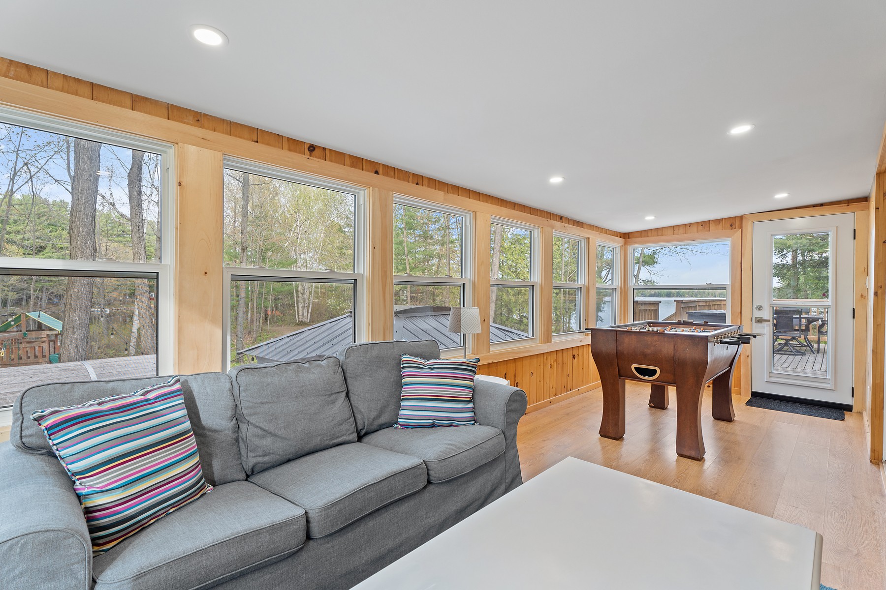 A sunroom with a big couch, lots of windows across a wood-panelled wall, and a fooseball table. A door leads out to a deck.