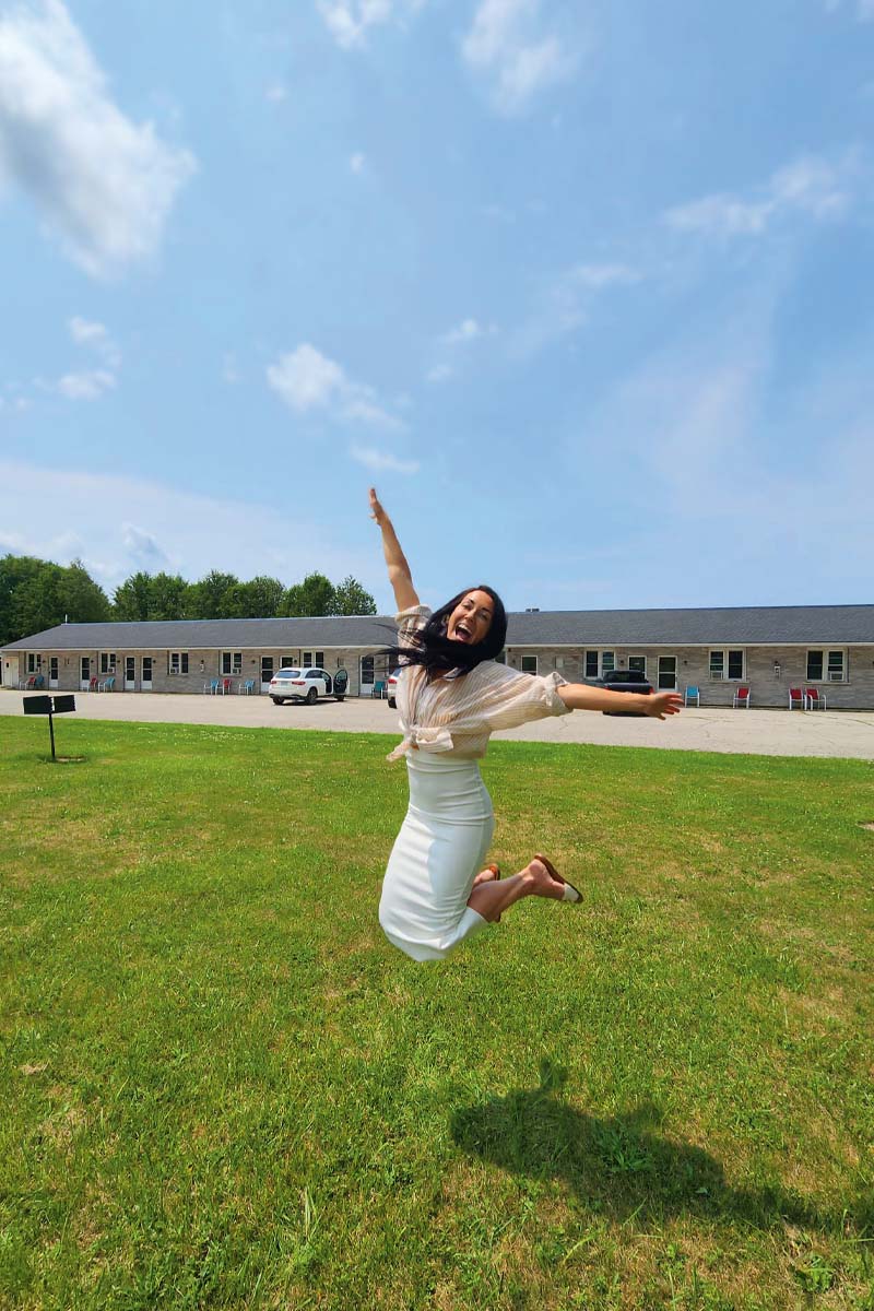 A woman in a white dress jumps in the area on a patch of grass in front of a long building