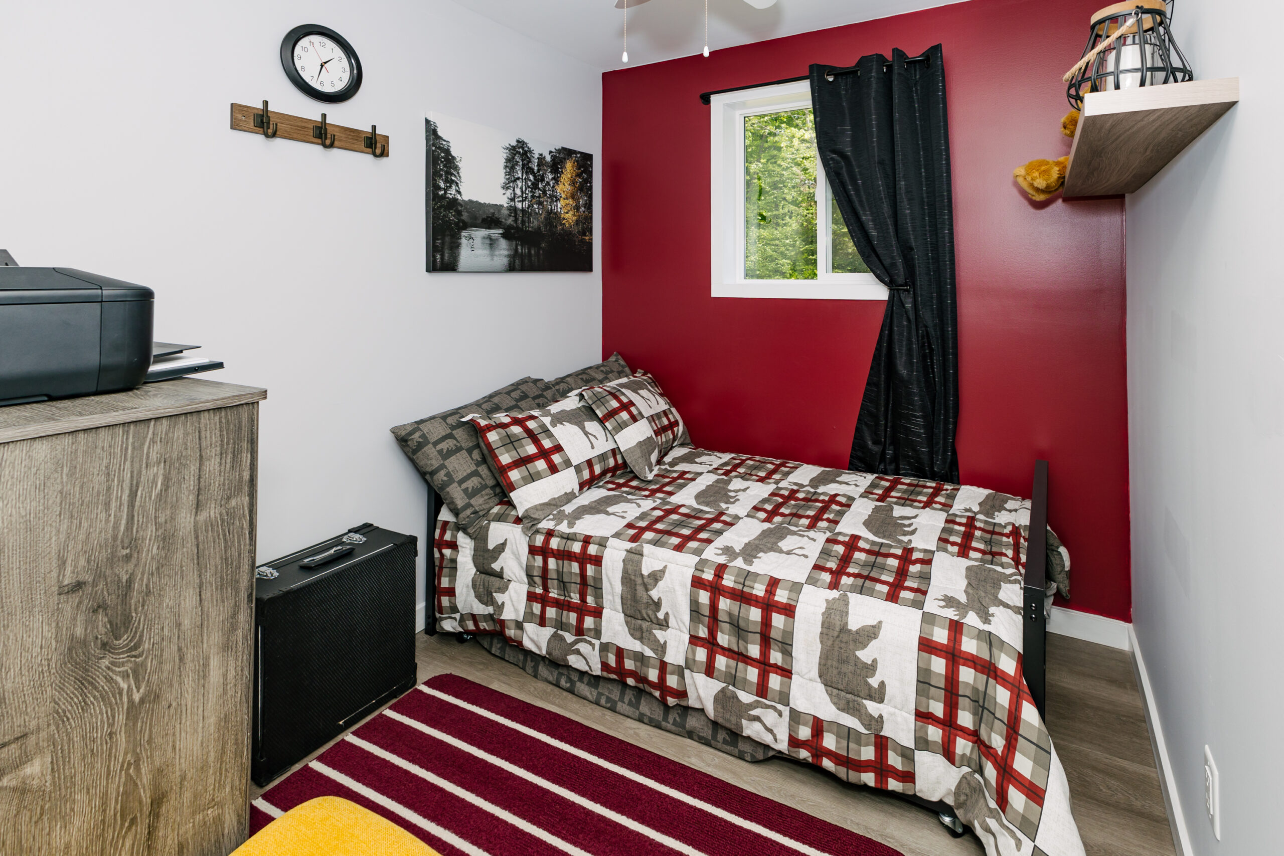 A small red and white bed pressed against a red wall with a window and a black curtain. On the ground, a red and white striped carpet