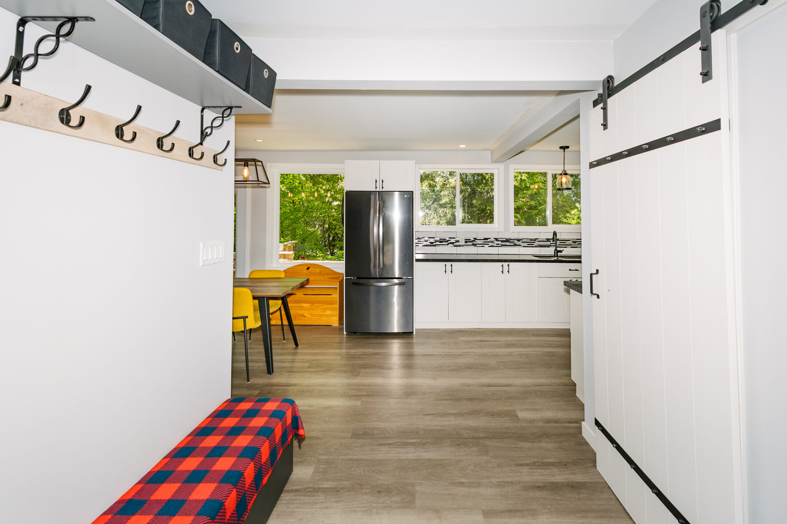 On the left of the entryway is a white wall with black hooks and a long black bench covered in a red and black blanket. The entrance faces a stainless steel fridge next to the kitchen counter