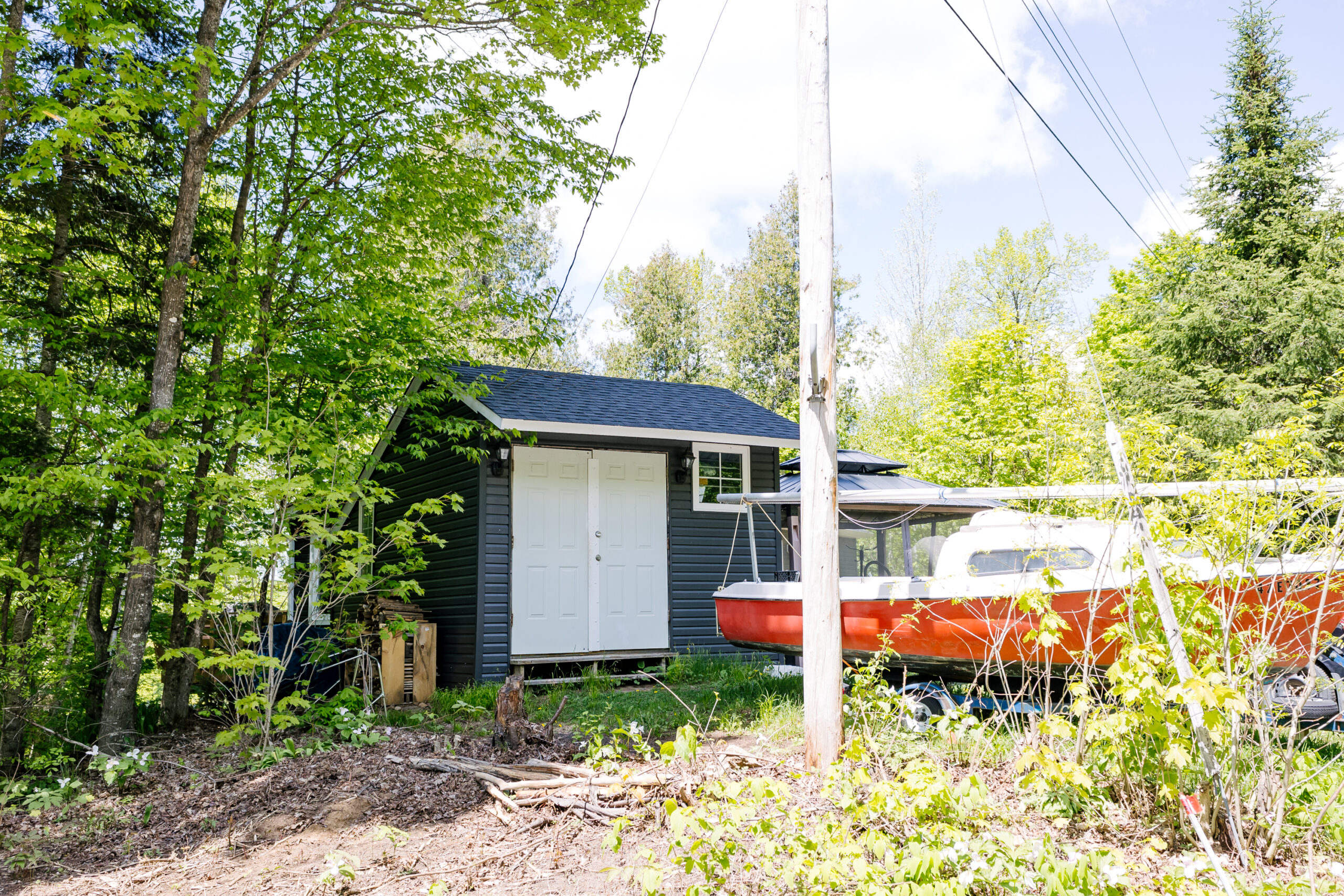 A blue-grey shed with white doors in the trees. A red and white boat sits on the grass beside it