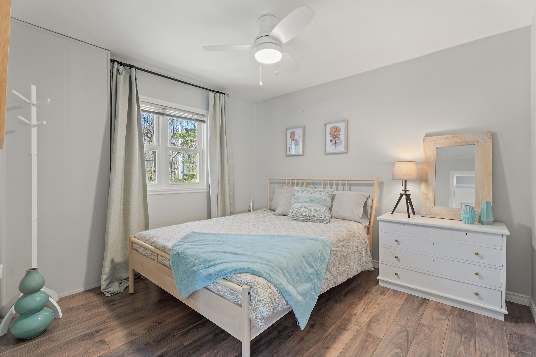 A white bed with a light blue blanket draped over the end next to a window and side table