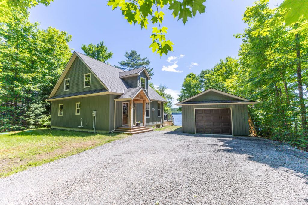 A green paneled cottage with a spacious gravel driveway and green freestanding garage