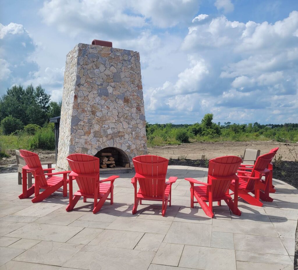A tall stone fire pit outdoors faces a half-moon of red Muskoka chairs