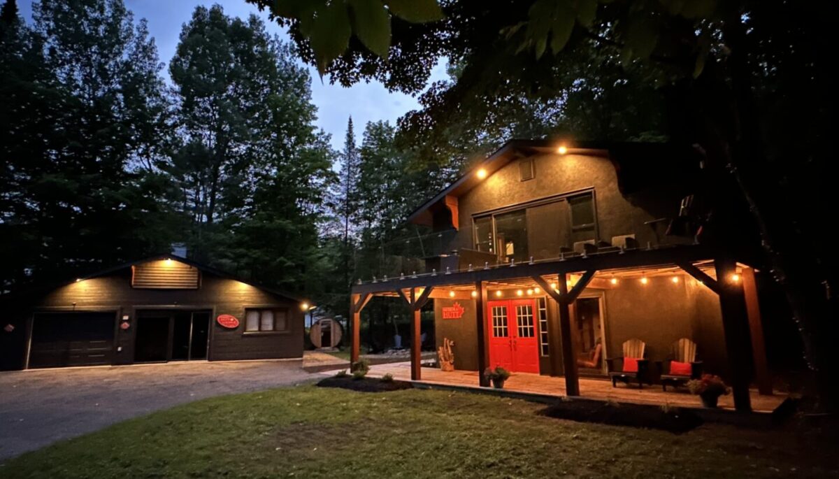 A wood cottage with red Muskoka chairs in the front is lit up by fairy lights at night
