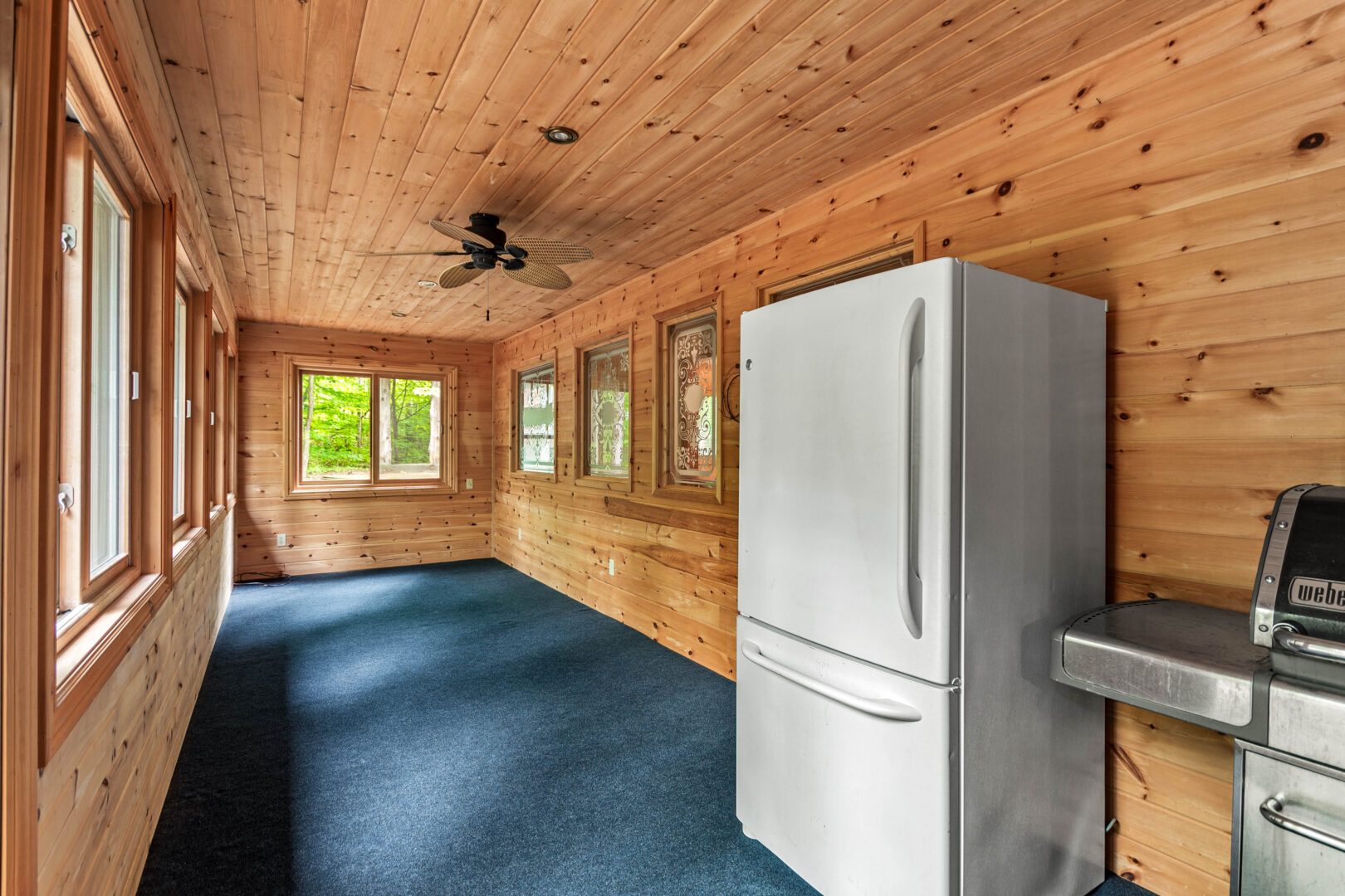 A wood paneled sunroom with blue carpeting and a white fridge